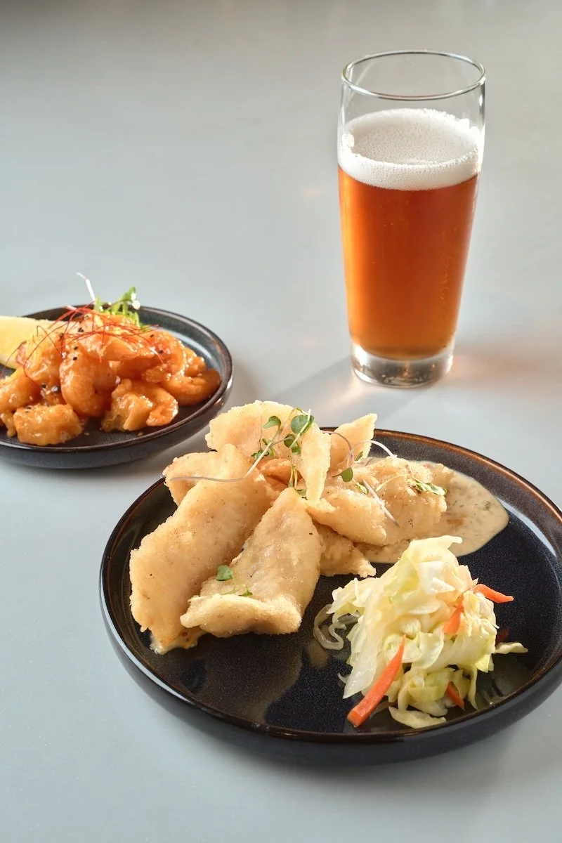 Fried fish fillets with coleslaw and a glass of amber beer on a light-colored table.