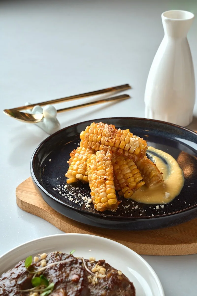 Fried corn on the cob served with a creamy sauce on a black plate, accompanied by a white pitcher and chopsticks in the background.
