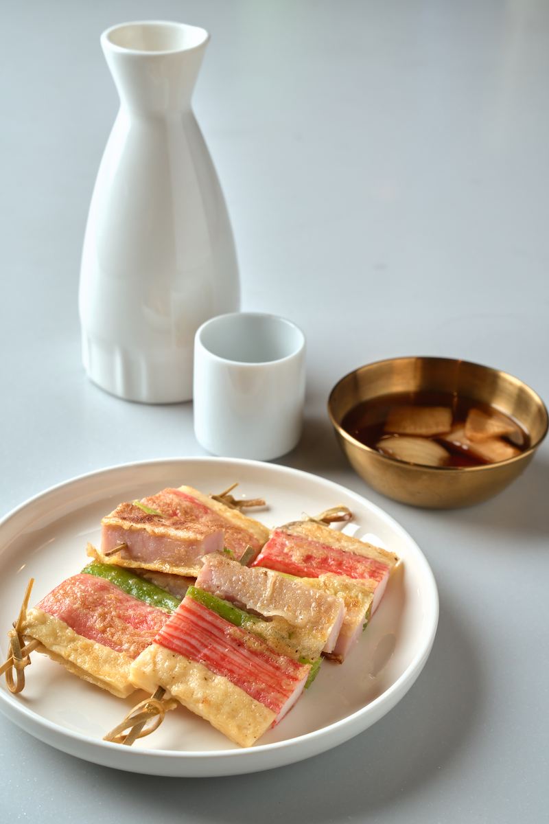 A white plate with Korean rice cakes (tteok) wrapped in fish or meat, tied with green onion stems, served with a bowl of soy sauce with garlic slices, a white ceramic pitcher, and a small white ceramic cup on a light gray surface.