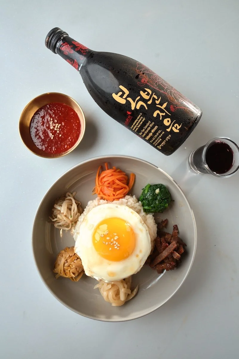 Bowl of Korean bibimbap with a fried egg on top, beside a bottle of Korean rice wine (soju), a small glass of soju, and side dishes of red sauce and dark beverage.