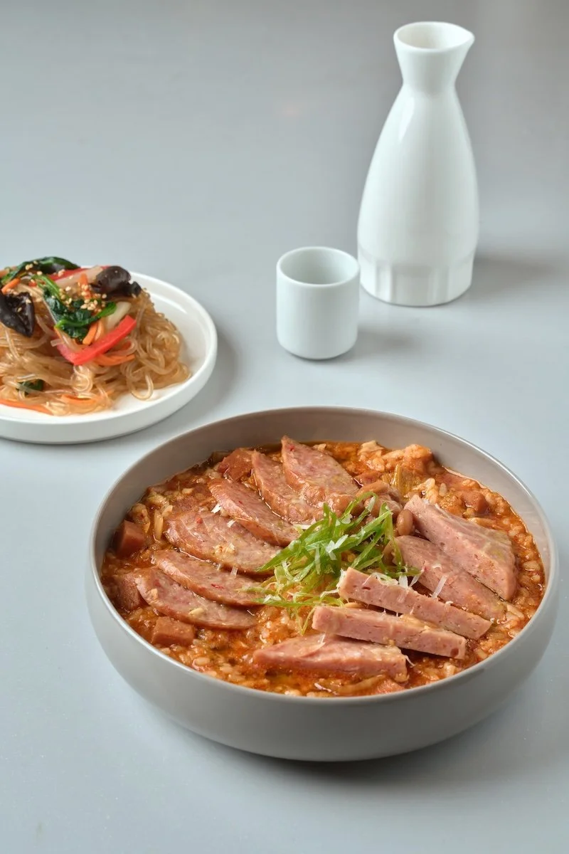 Bowl of rice with sliced roasted pork, garnished with green onions, on a table with a side of noodle salad, a white carafe, and a small white cup, against a plain gray background.