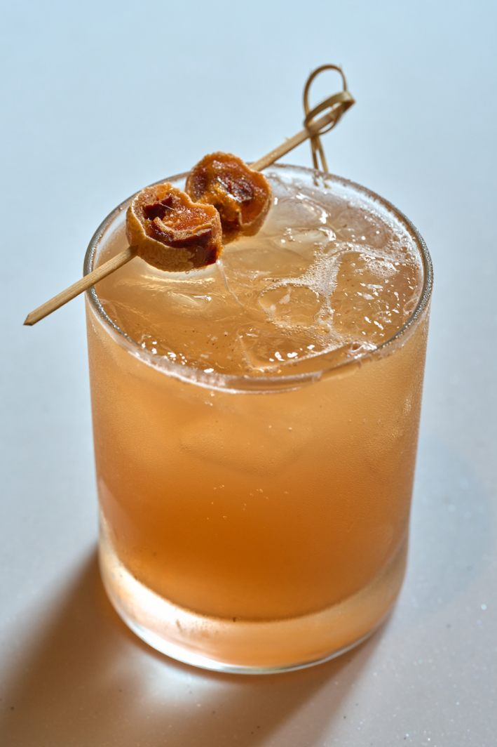 A golden-brown traditional Korean cinnamon punch with ice, topped with a skewer of dried fruit, presented in a clear glass on a soft, minimal background.