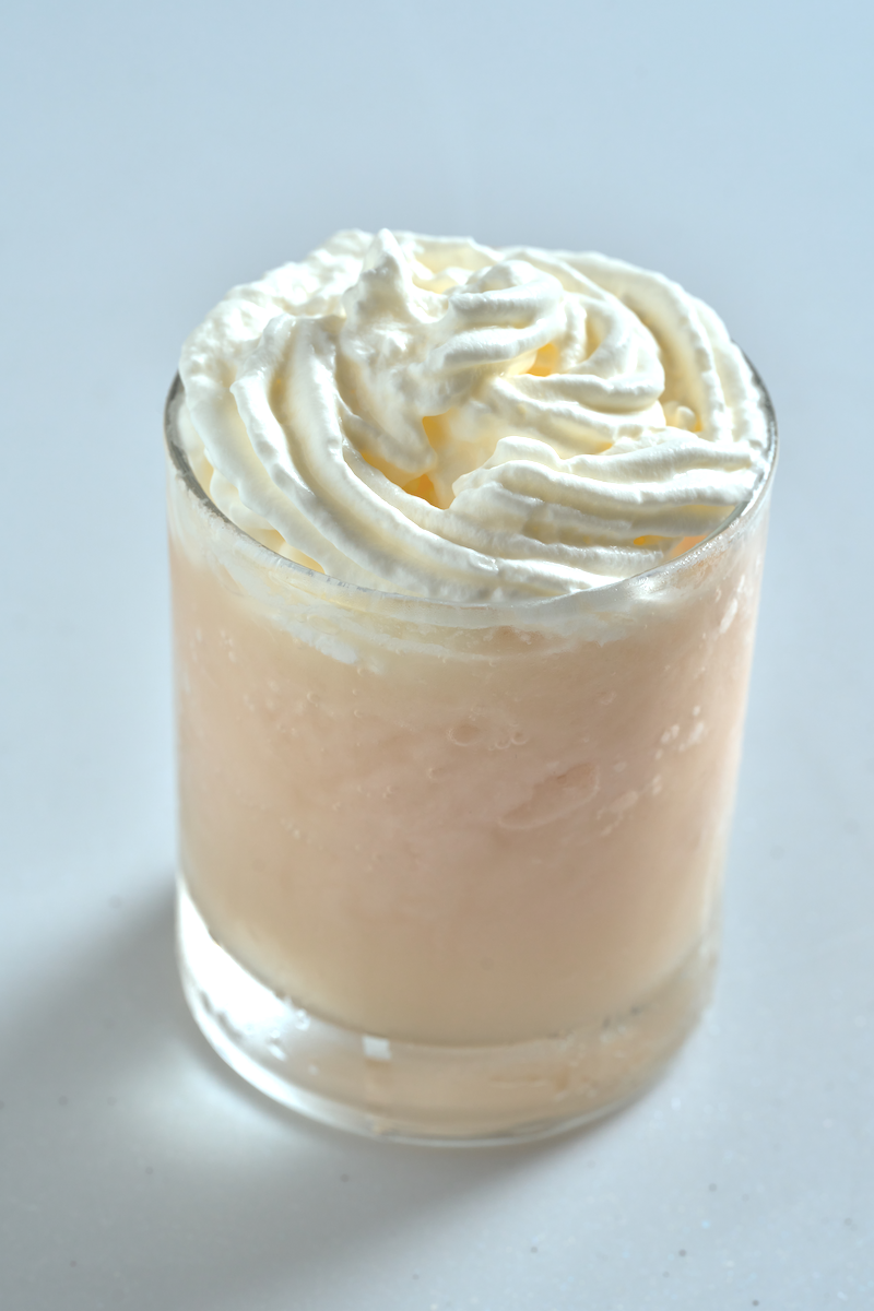 A pale pink frozen lychee makgeolli topped with whipped cream, presented in a clear glass on a neutral backdrop.
