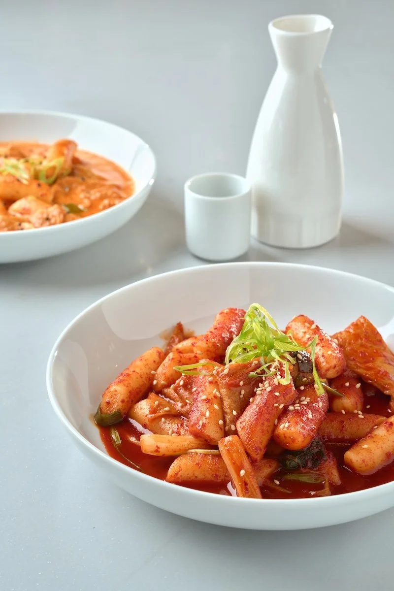 A white bowl of Korean spicy rice cake (tteokbokki) garnished with green onions and sesame seeds, with a background of another bowl of food, a white ceramic pitcher, and a small cup on a light-colored surface.