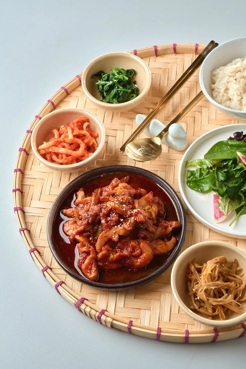Traditional Korean meal with rice, kimchi, seasoned greens, marinated vegetables, and a spicy pork dish on a woven tray.