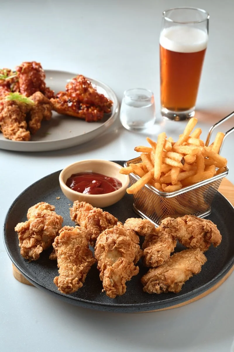 Fried chicken wings with a side of French fries and ketchup, a glass of beer, and a plate of fried chicken with sauce in the background.