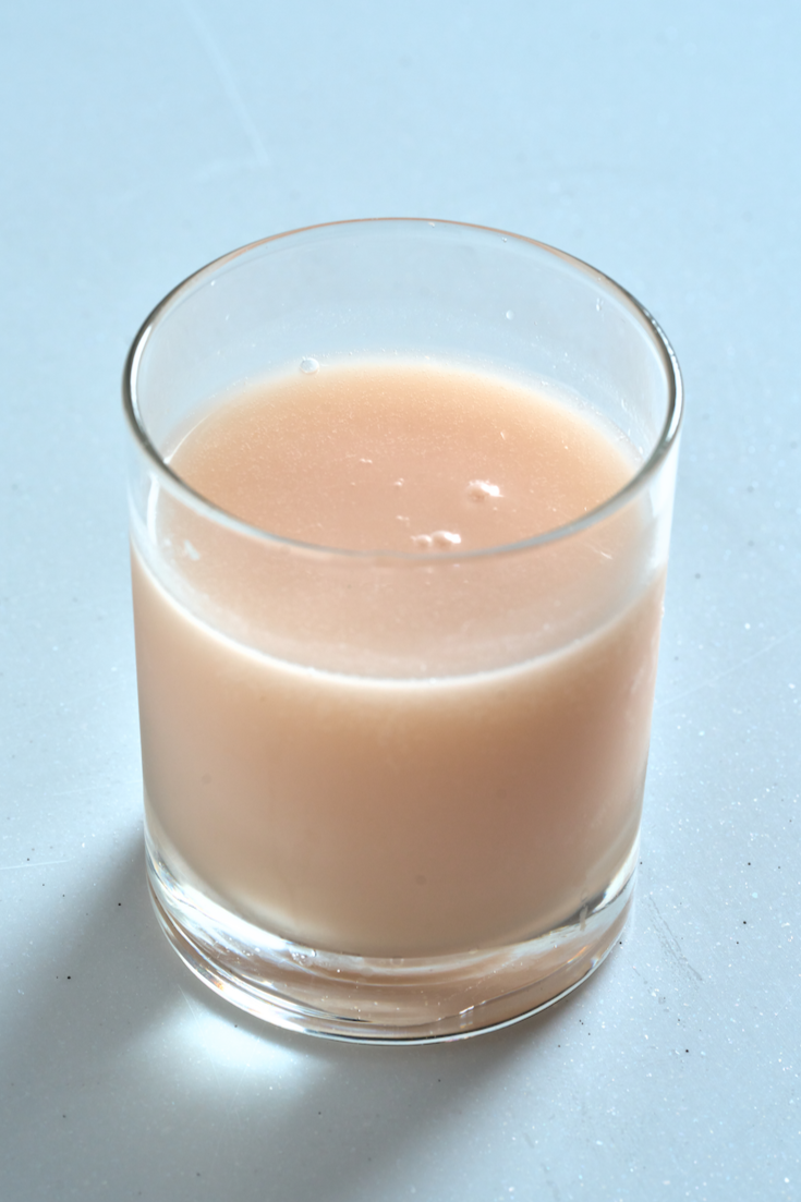 A pale pink, creamy lychee makgeolli in a clear glass, smooth and opaque, set against a light, modern background.