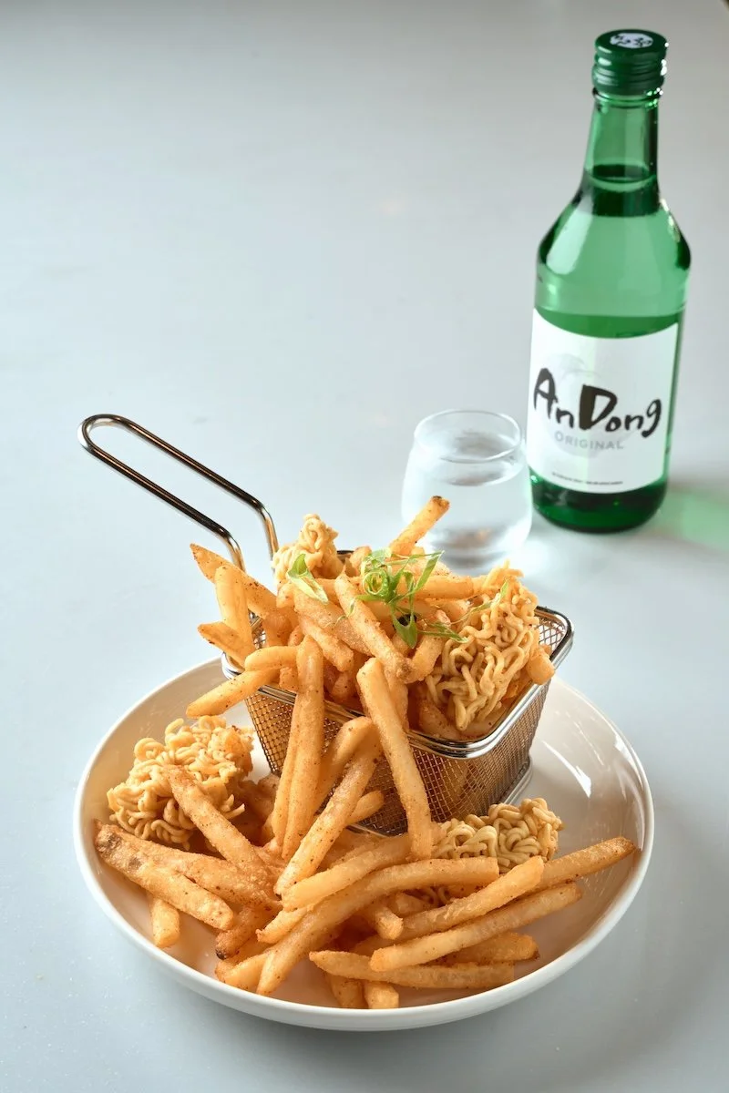 Fried seasonings and noodles served with French fries, a glass of water, and a bottle of Soju on a white surface.