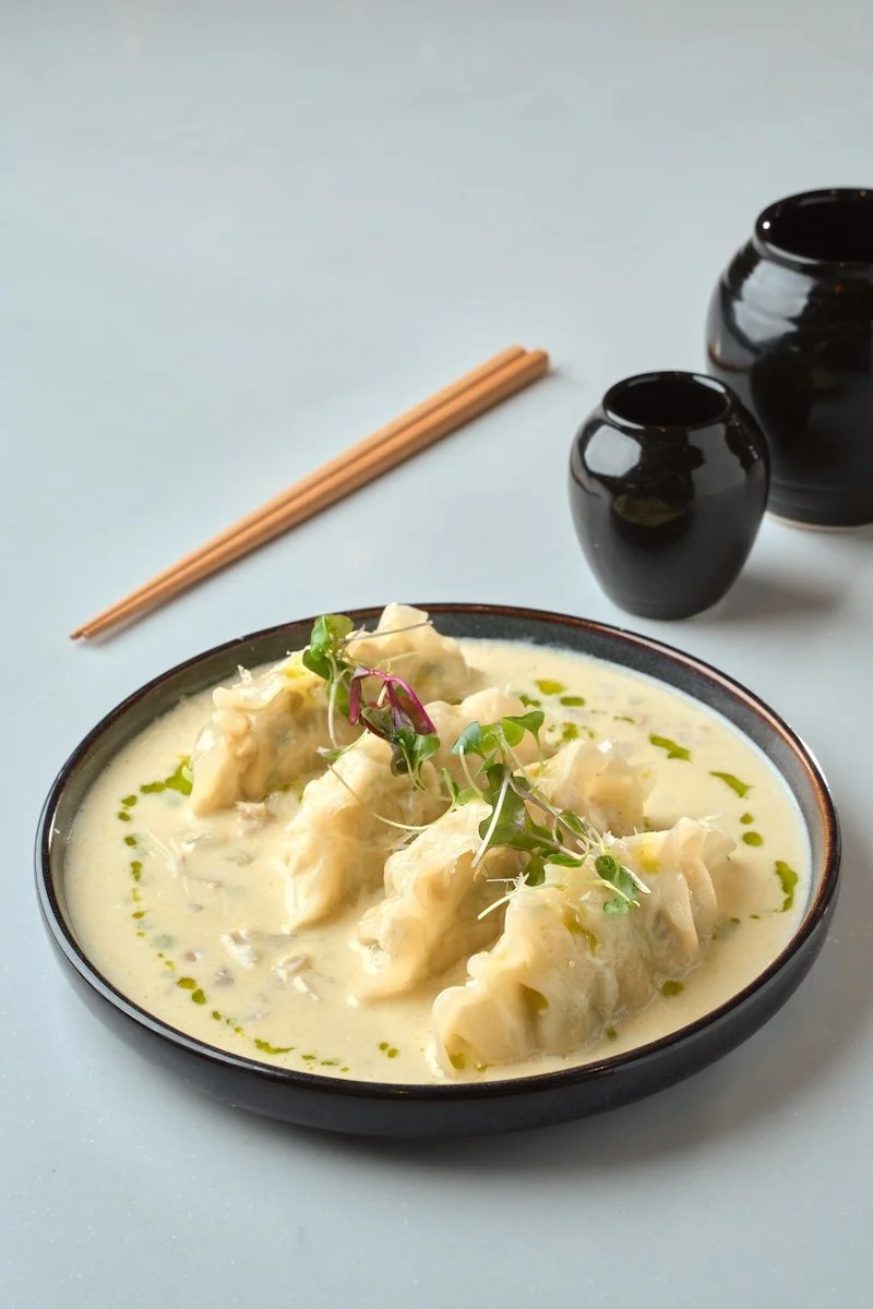 A bowl of Asian dumplings in a creamy sauce garnished with microgreens, with two black ceramic vases and a pair of wooden chopsticks in the background.