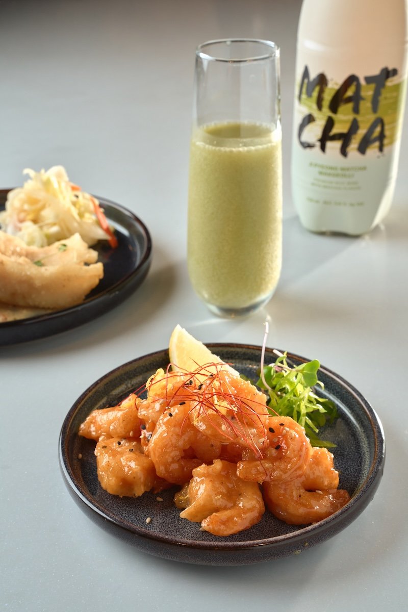 Cooked shrimp coated in sauce served with a lemon wedge and microgreens on a dark plate, with a side of food on a black dish and a green fruit smoothie in a tall glass in the background.