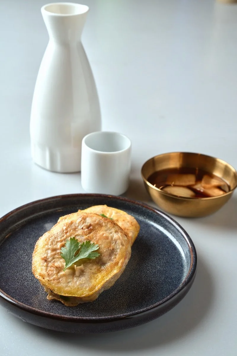 Fried eggplant slices garnished with a cilantro leaf on a dark ceramic plate, accompanied by a bowl of dipping sauce, a white ceramic vase, a small white cup, and a gold-colored bowl of soy sauce on a white surface.