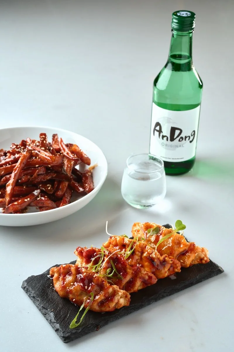 A dish of Korean fried chicken with sauce and garnish on a black slate plate, a bowl of stir-fried beef with sesame seeds, a small glass of clear beverage, a bottle of Soju, and a glass of clear liquid on a white surface.
