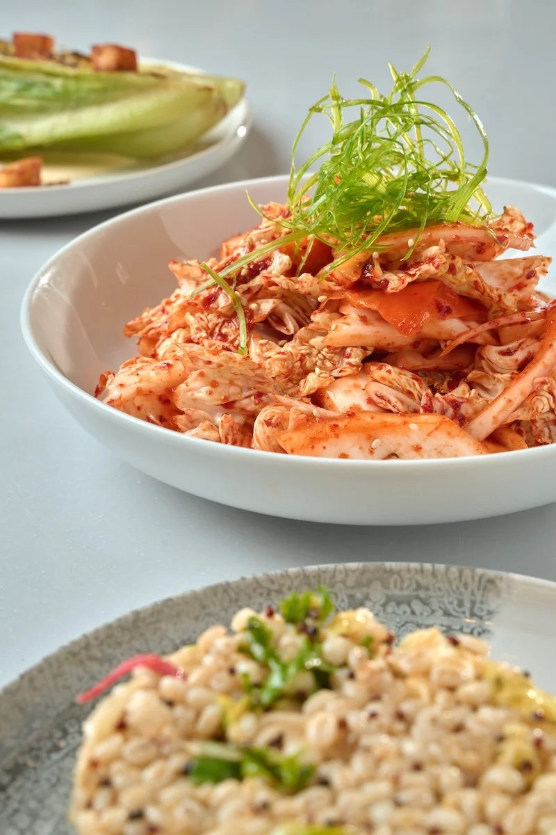 Bowl of kimchi garnished with green shredded vegetables, with other dishes partially visible in the background