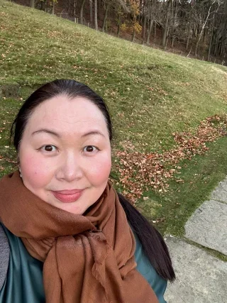 A woman with dark hair and light skin is smiling outdoors, wearing a brown scarf and teal jacket, with a grassy area and trees in the background.