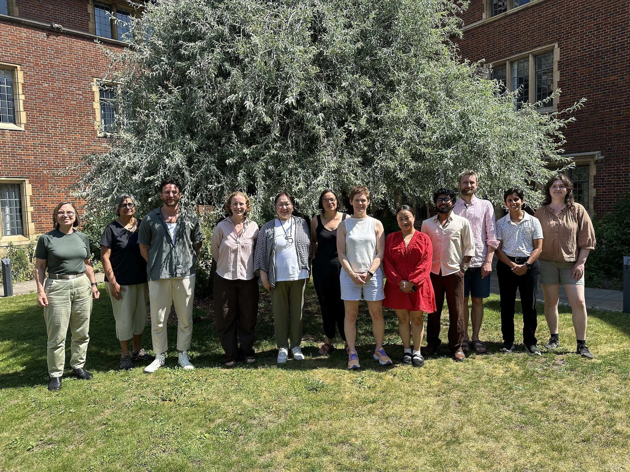 A group of fifteen people standing outside on grass in front of a large leafy bush, with brick buildings and windows in the background.