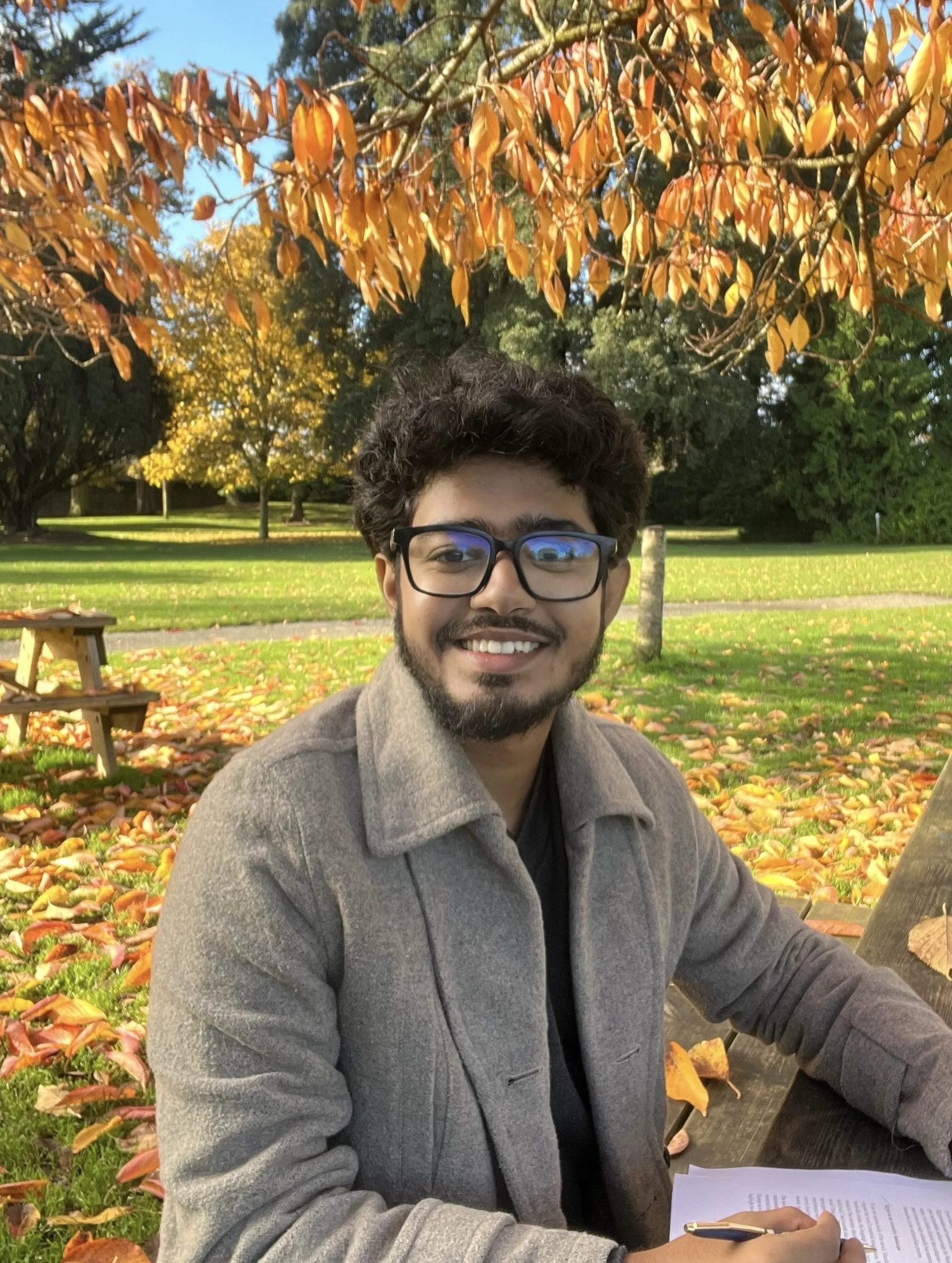A man with curly hair and glasses smiling outdoors in a park with trees and yellow-orange leaves, sitting at a table with papers and a pen.
