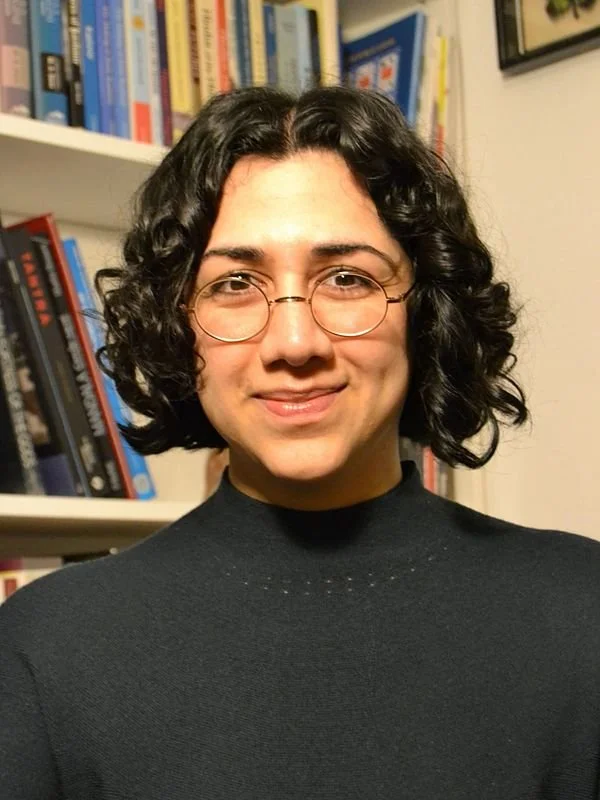 A woman with curly dark hair and glasses smiling in front of a bookshelf filled with books.