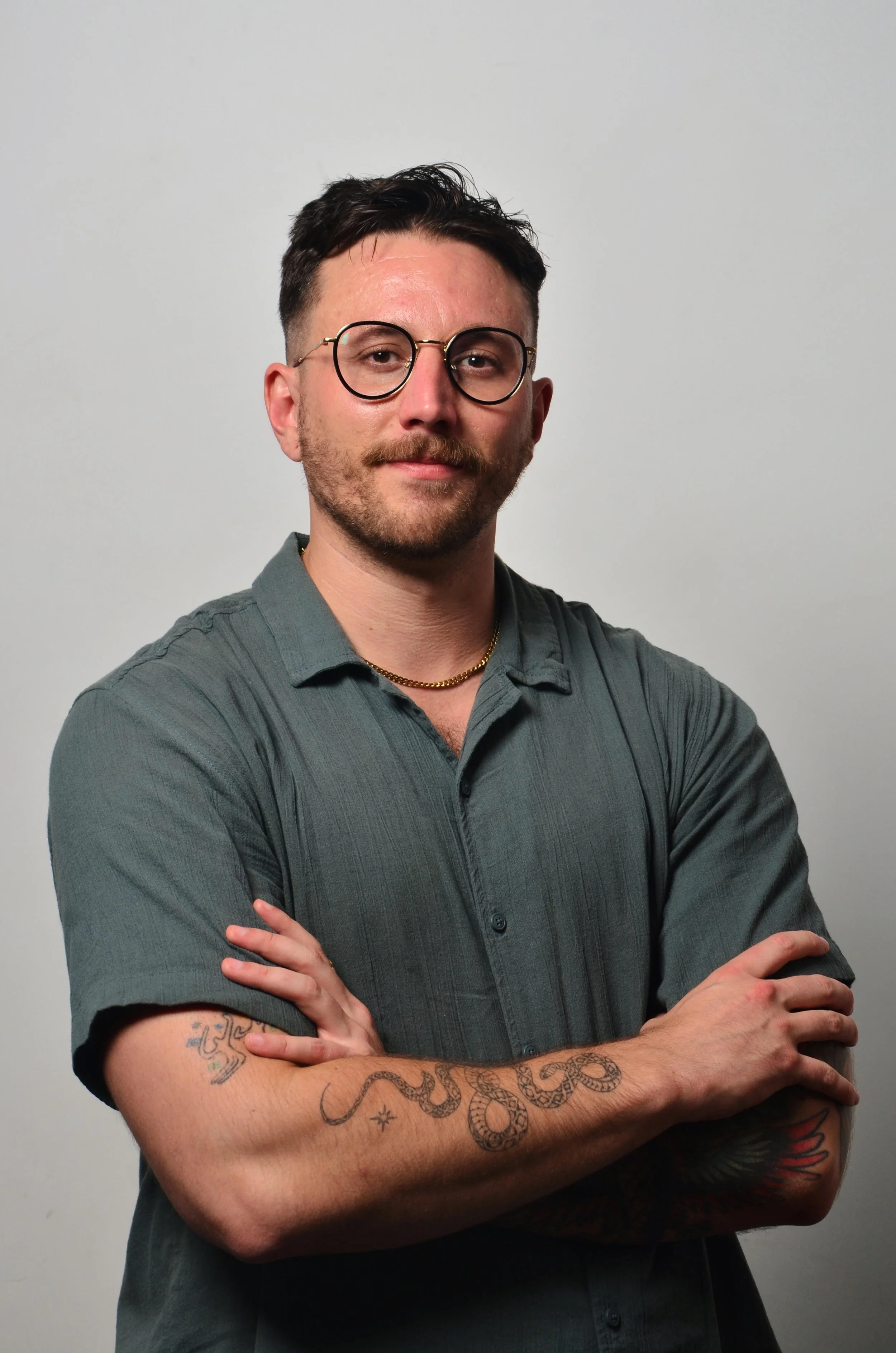 A man with short dark hair and glasses, wearing a gray shirt, standing with arms crossed against a plain gray background.