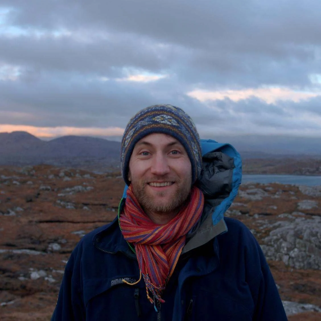 A man in outdoor gear smiling in a mountainous landscape with cloudy skies, hills, and a body of water in the background.