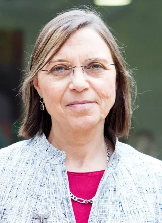 Portrait of a middle-aged woman with short gray hair and glasses, outdoors with greenery in the background.