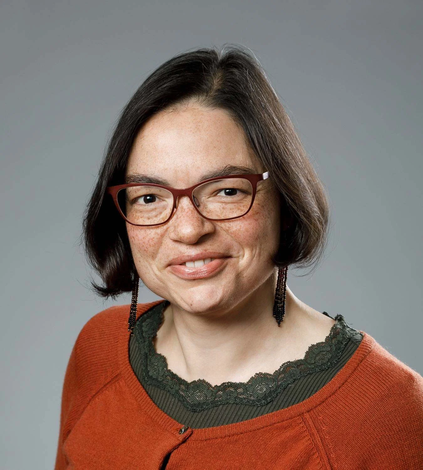 Portrait of a woman with short dark hair, wearing red glasses, earrings, an orange sweater, and a black lace-trimmed top, smiling against a gray background.