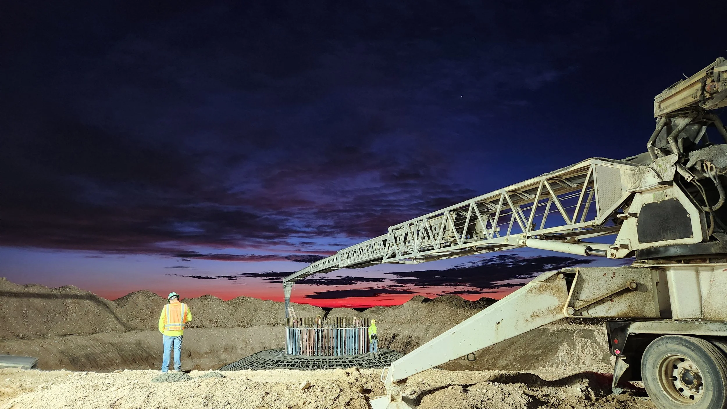Construction workers at a site with large machinery during sunset, featuring a crane and mounds of dirt.