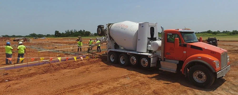 Construction site with workers in bright yellow safety vests and helmets pouring concrete, with a large orange cement mixer truck nearby.