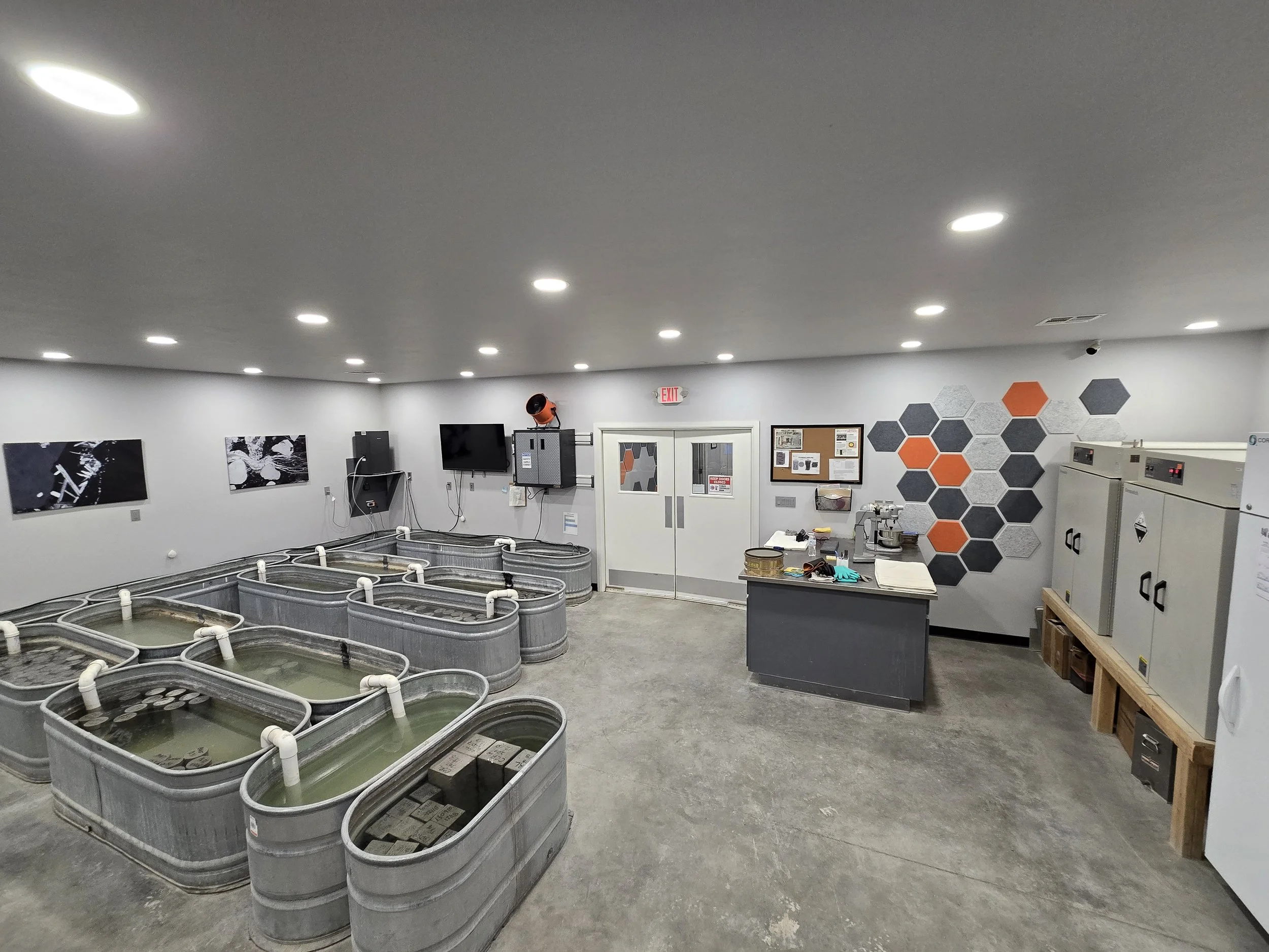 A room with multiple vats filled with a liquid, possibly in a laboratory or industrial setting. The room has white walls, bright ceiling lights, and various equipment on tables and walls, including a decorated wall with hexagonal panels.