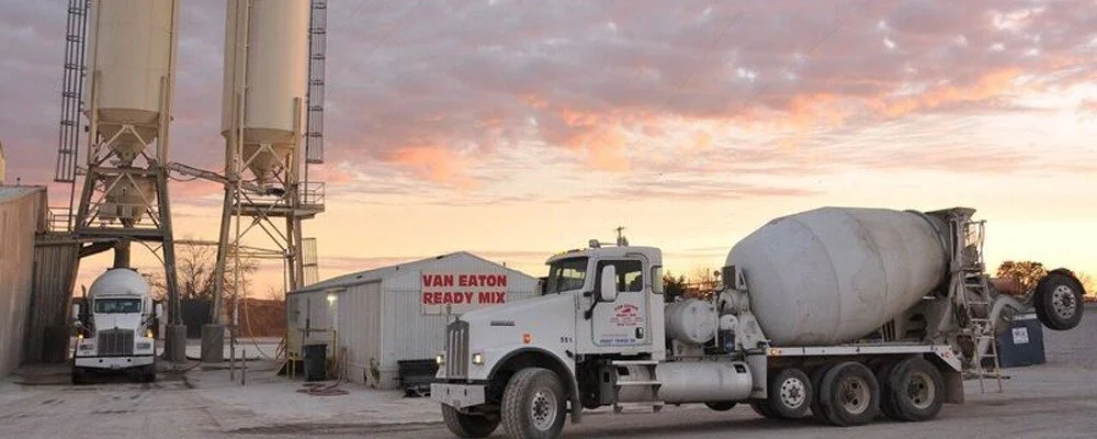 Concrete mixer truck parked at a construction site during sunset, with silos and a truck labeled 'Van Eaton Ready Mix' nearby.