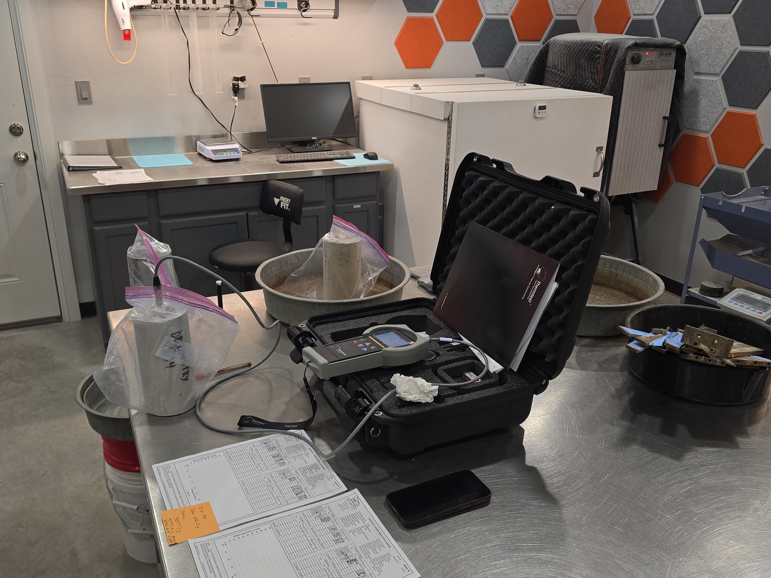 A cluttered laboratory or workshop space with electronic equipment, papers, and containers on a table. Various tools and devices are visible, including a monitor, a scale, and testing instruments.