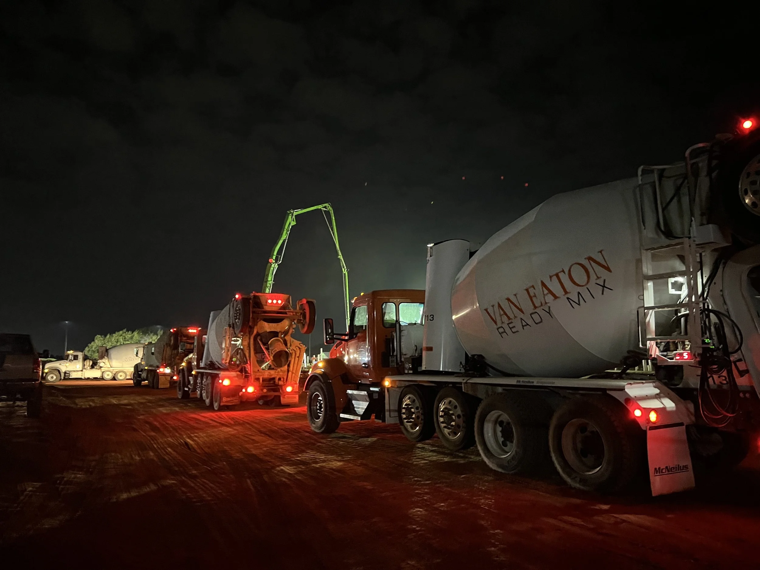 Nighttime view of cement mixers and construction vehicles parked outdoors, with a dark sky and cloudy weather. The scene is illuminated by artificial lighting.