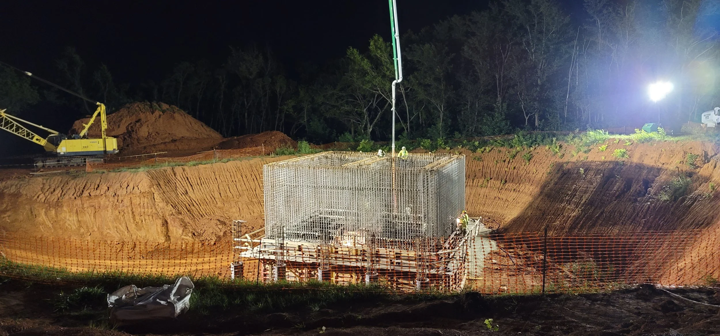 Nighttime construction site with workers and equipment, including a deep excavation, scaffolding structure, a crane, and large floodlights illuminating the area, surrounded by trees.