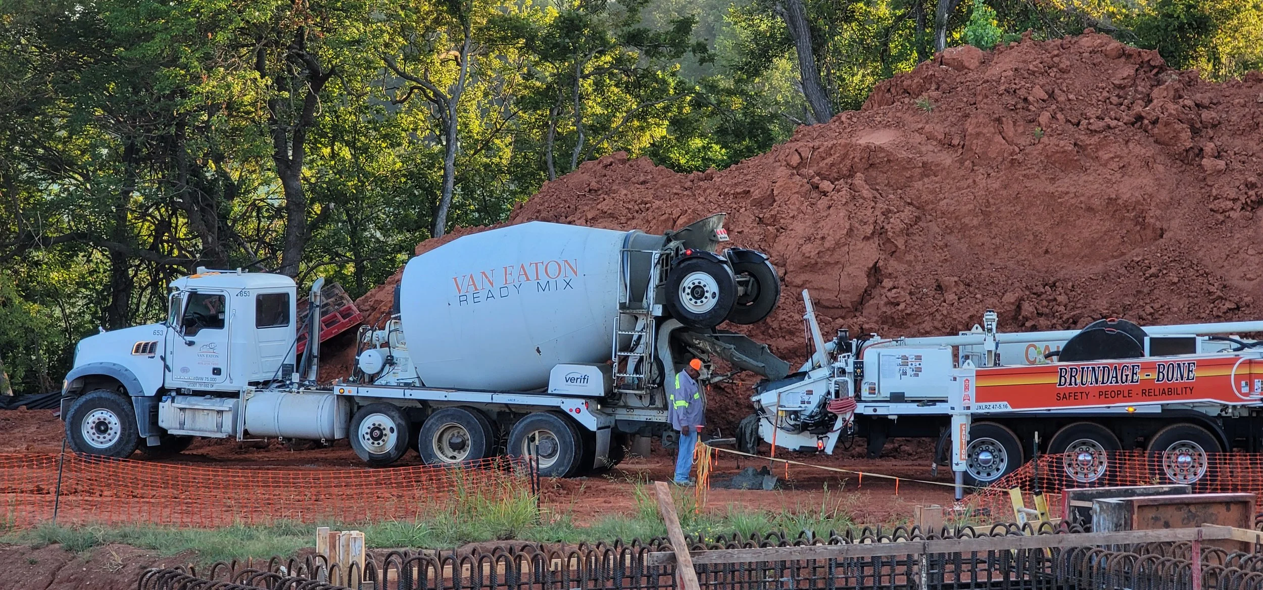 A construction site with a large cement mixer truck and a concrete pump with brand names Van Eaton Ready Mix and Brundage Bone. Two workers wearing safety vests and orange helmets work near a large pile of reddish dirt, with a green wooded background.