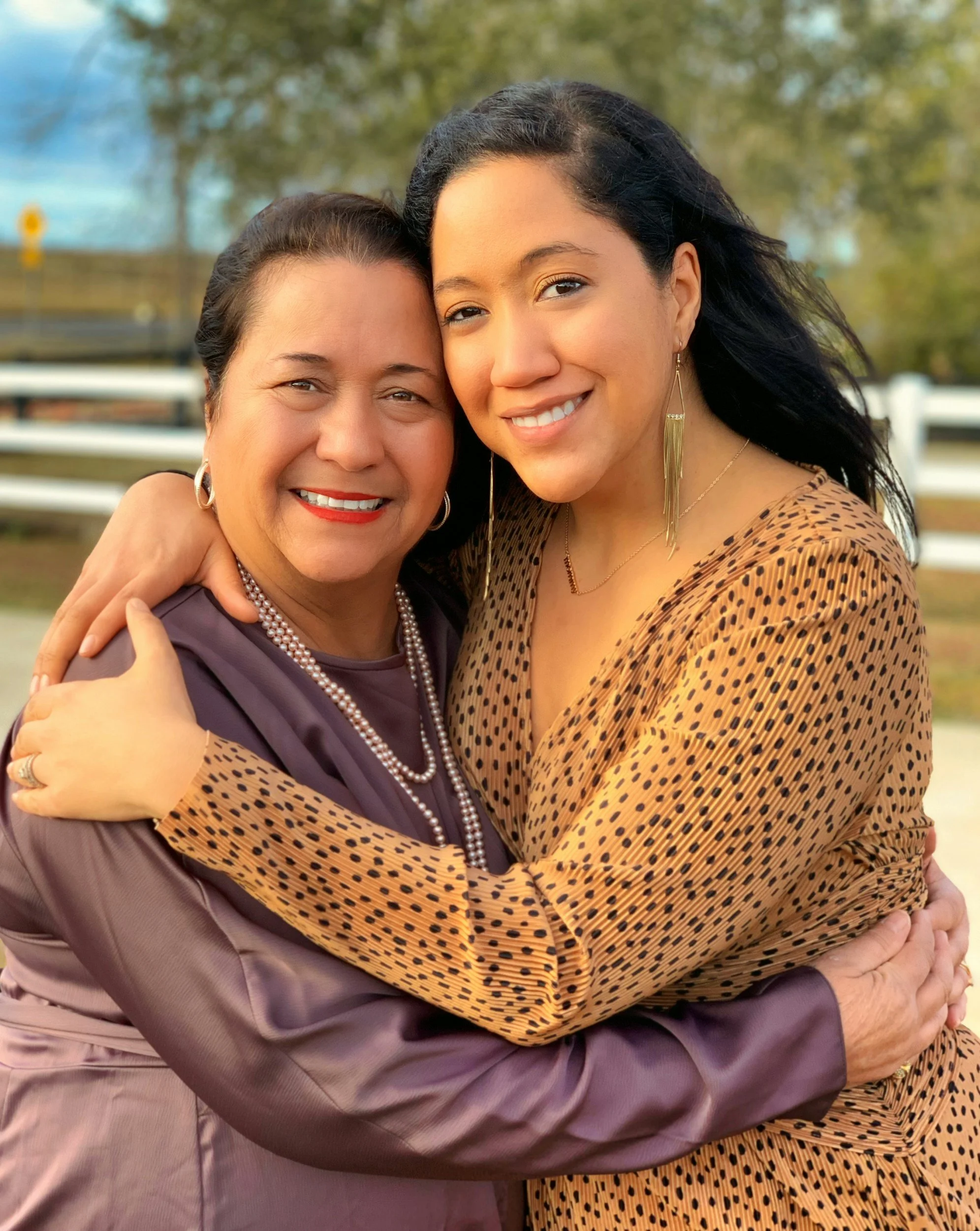 Two women hugging outdoors, smiling at the camera, with trees and a white fence in the background.