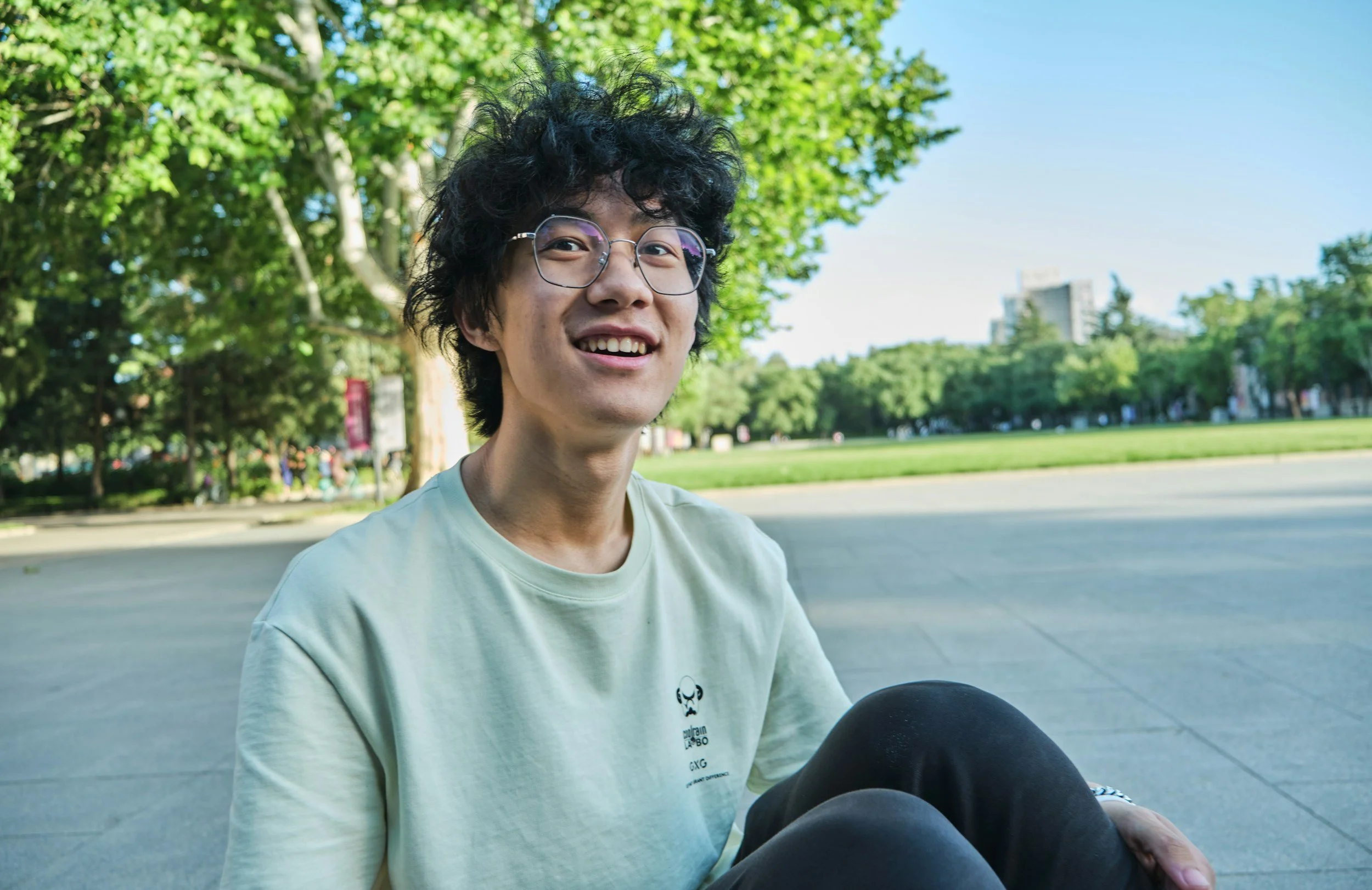 A young man with curly black hair and glasses smiling while sitting outdoors in a park with green trees and a blue sky in the background.