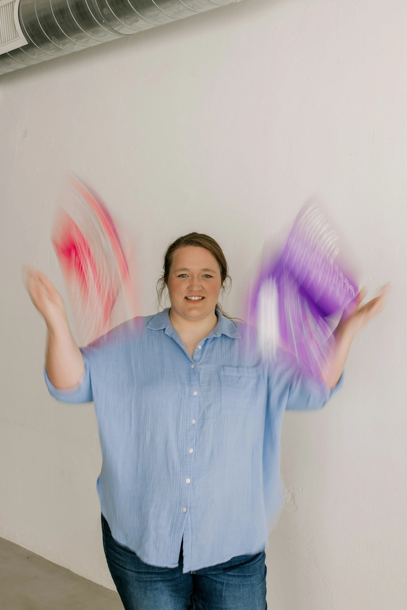 A woman in a blue shirt and jeans is smiling while waving her arms, with blurred motion of pink and purple fabric or objects around her hands, against a plain white wall background.