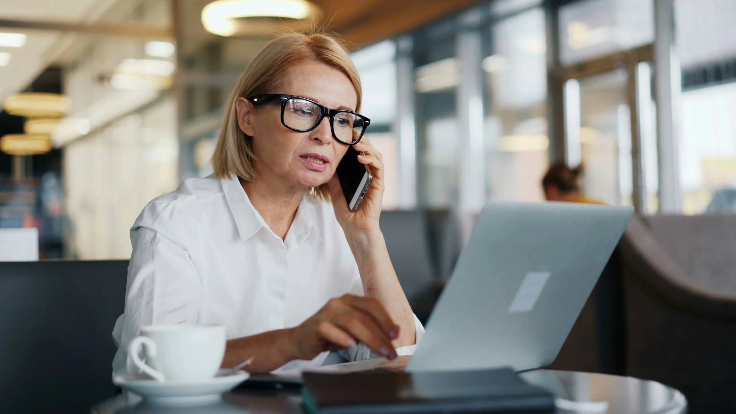 A middle-aged woman with blonde hair, wearing glasses and a white shirt, is sitting at a table in a modern coffee shop or café. She is talking on a cell phone while working on a silver laptop. There is a white coffee mug and a smartphone on the table in front of her. The background shows large windows and a softly lit interior.
