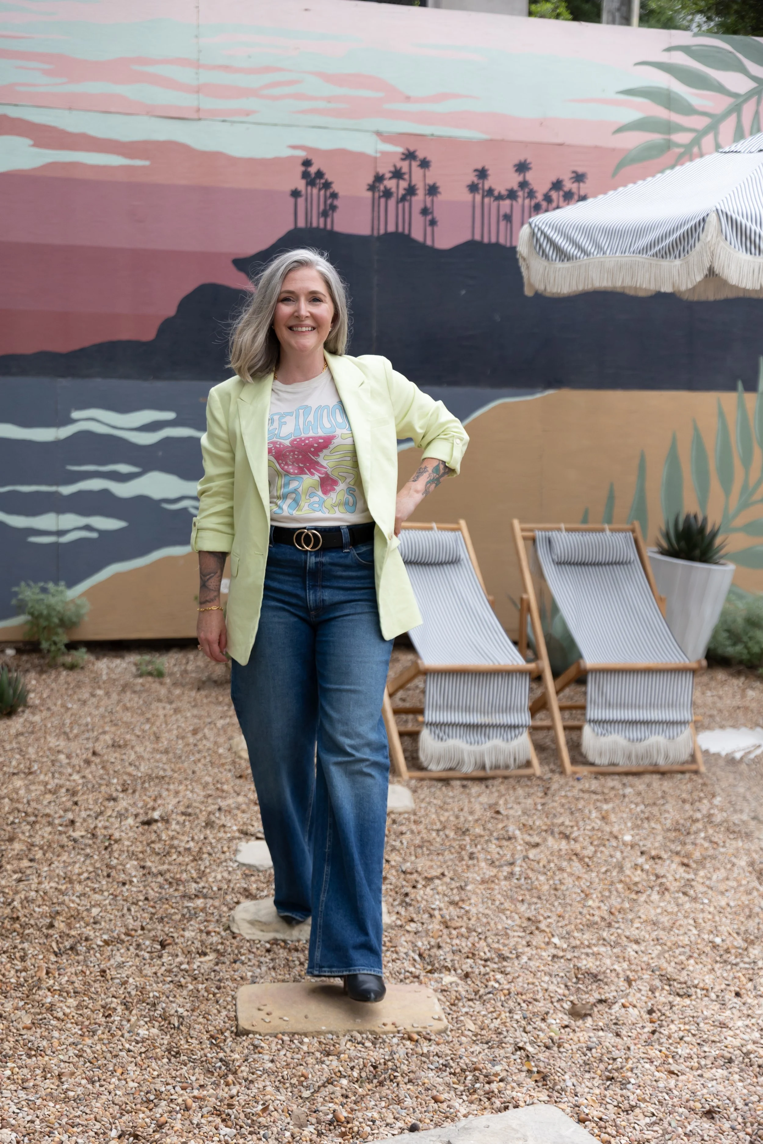 Smiling woman with long gray hair, wearing a light yellow blazer, graphic t-shirt, and blue jeans, standing on a stepping stone in an outdoor area with a colorful mural of palm trees and mountains in the background. There are three striped deck chairs and a large planter with a succulent plant beside her, with a striped umbrella overhead.