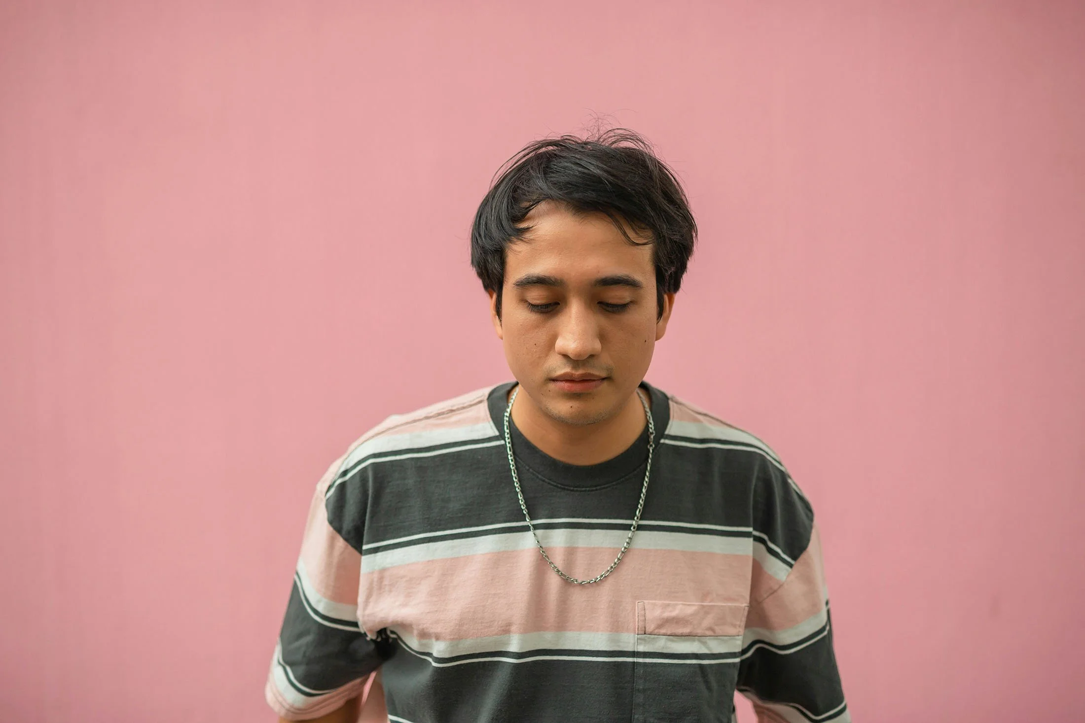 A young man with short dark hair, closed eyes, and light skin, wearing a striped t-shirt and a silver chain necklace, standing against a pink background.
