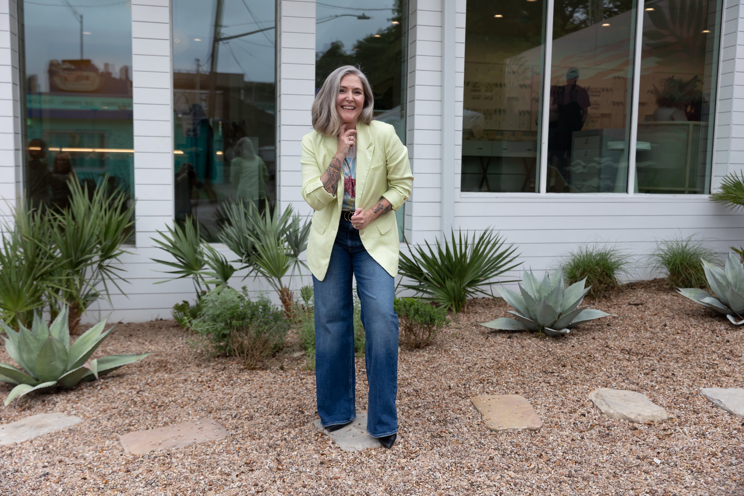 A smiling woman with shoulder-length gray hair and tattoos on her arms standing outside in front of a white building, wearing a light yellow blazer, graphic T-shirt, and blue jeans, surrounded by desert plants and gravel.