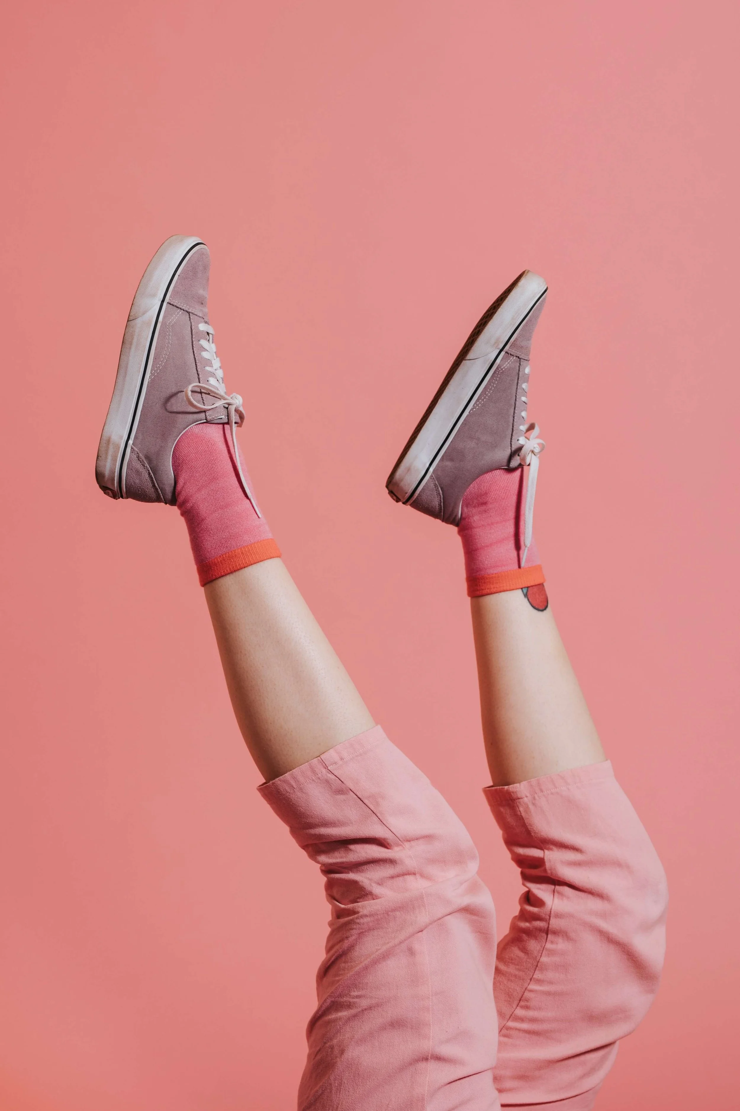 Close-up of legs in pink pants and pink sneakers with white soles and laces, raised against a pink background.