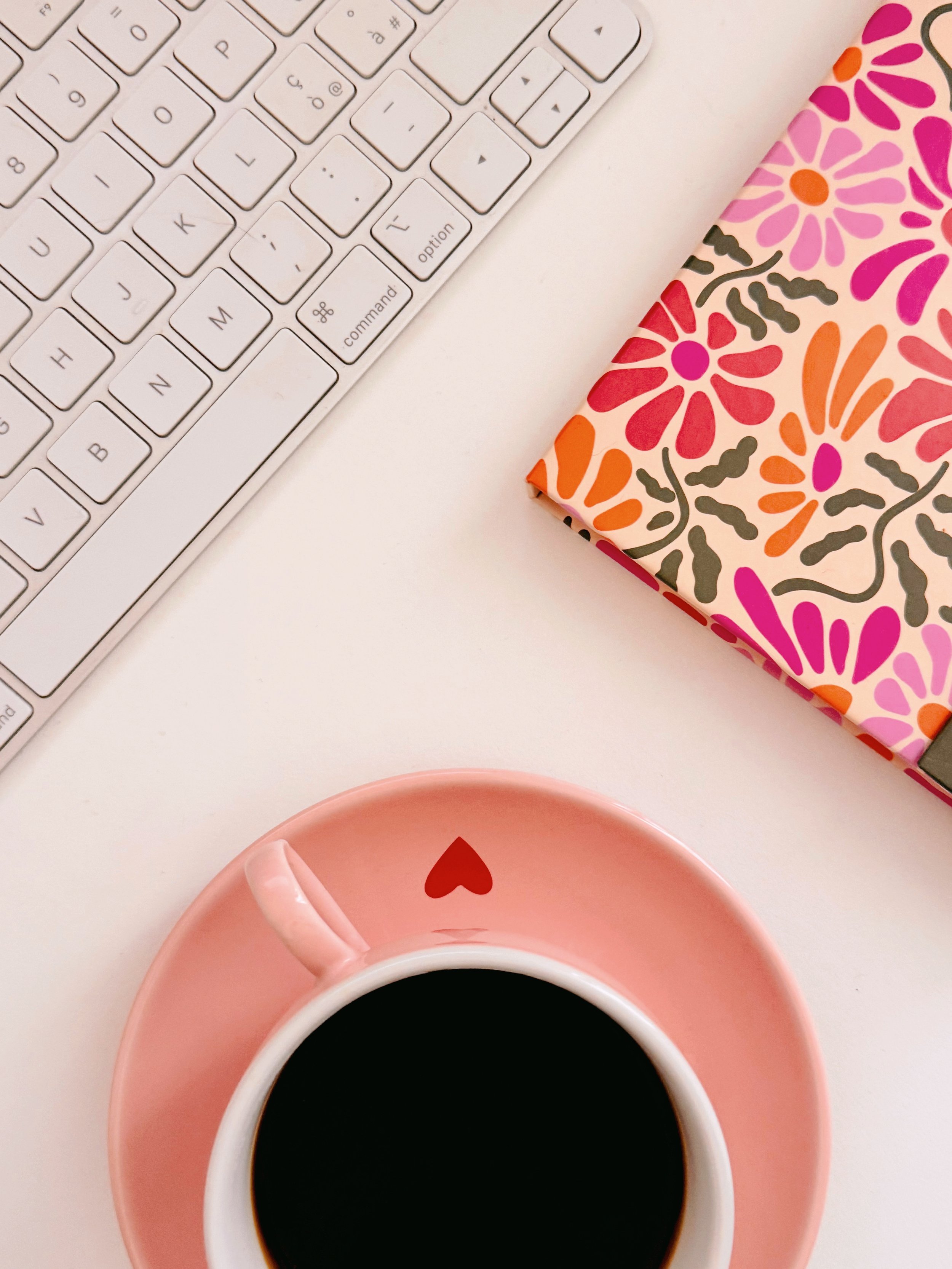 White keyboard with a pink floral notebook and a pink cup of black coffee on a white desk.