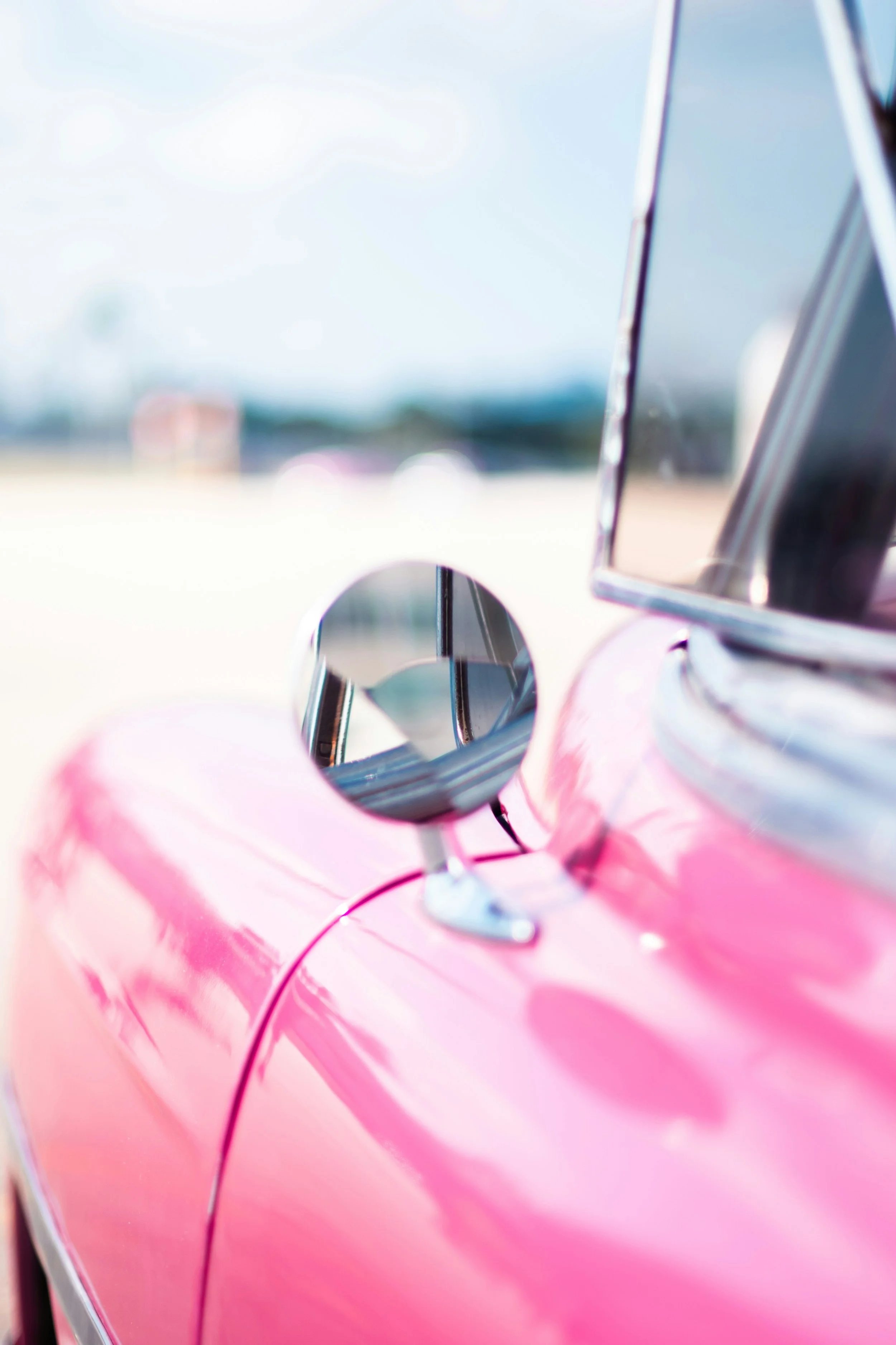 Close-up of the side mirror and door of a pink vintage car with chrome details, parked outdoors.