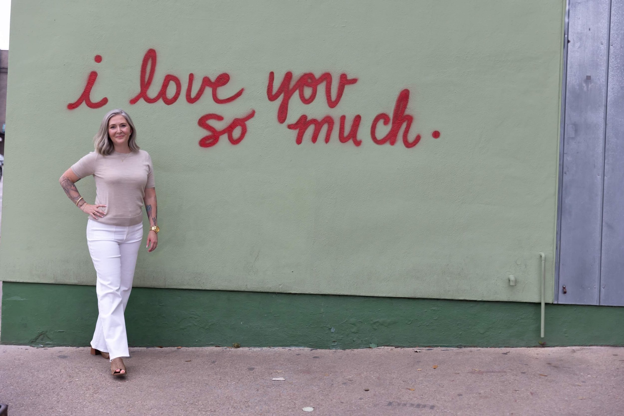A woman standing in front of a green wall with red graffiti that says "I love you so much." She is wearing a beige top and white pants, with her hand on her hip, smiling at the camera.