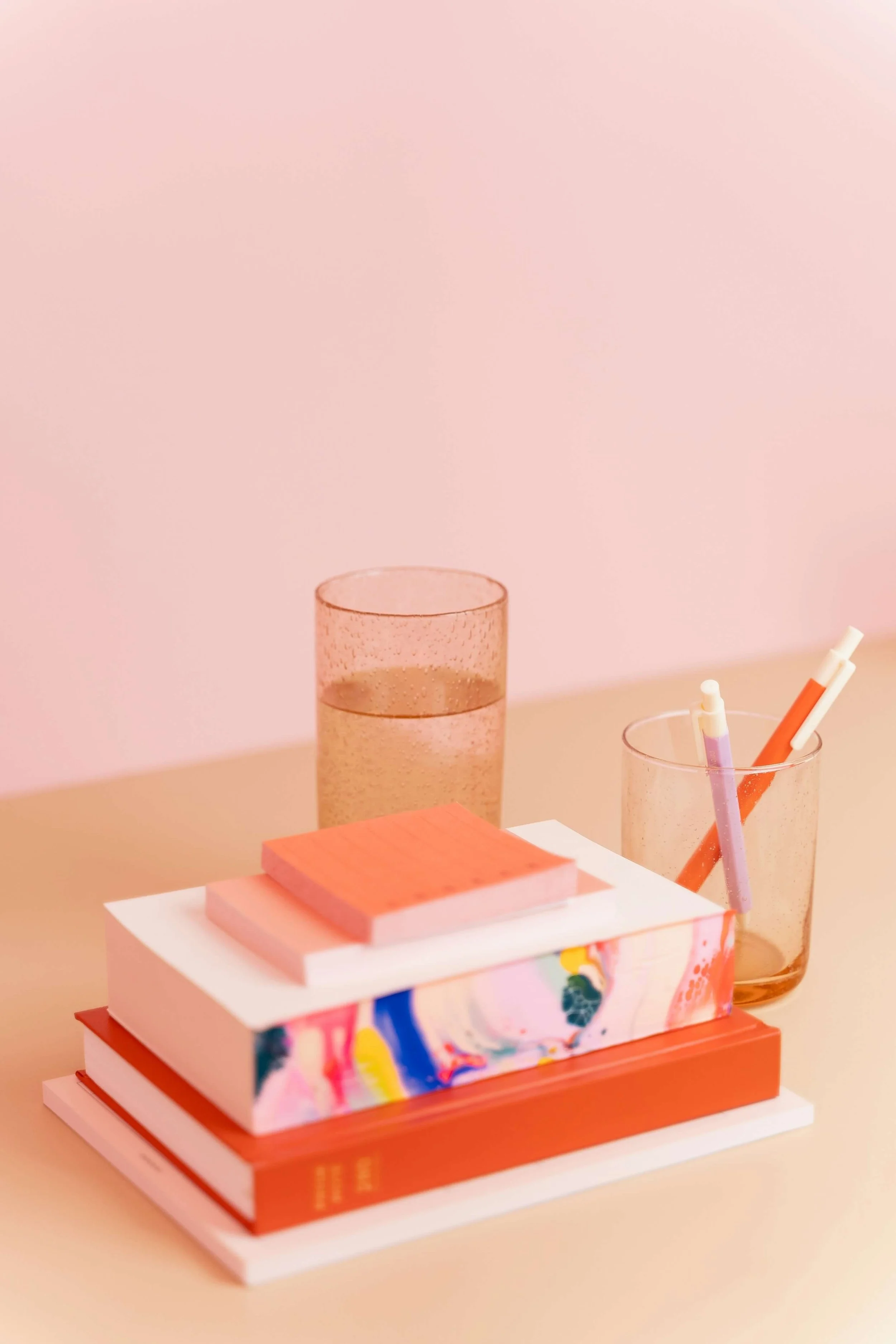 A stack of colorful books on a white table, with a small white sculpture or decorative object on top. Behind the books, there are two pink-tinted glass containers; one is empty, and the other contains a few colored pens or markers. The background is a plain, soft pink wall.