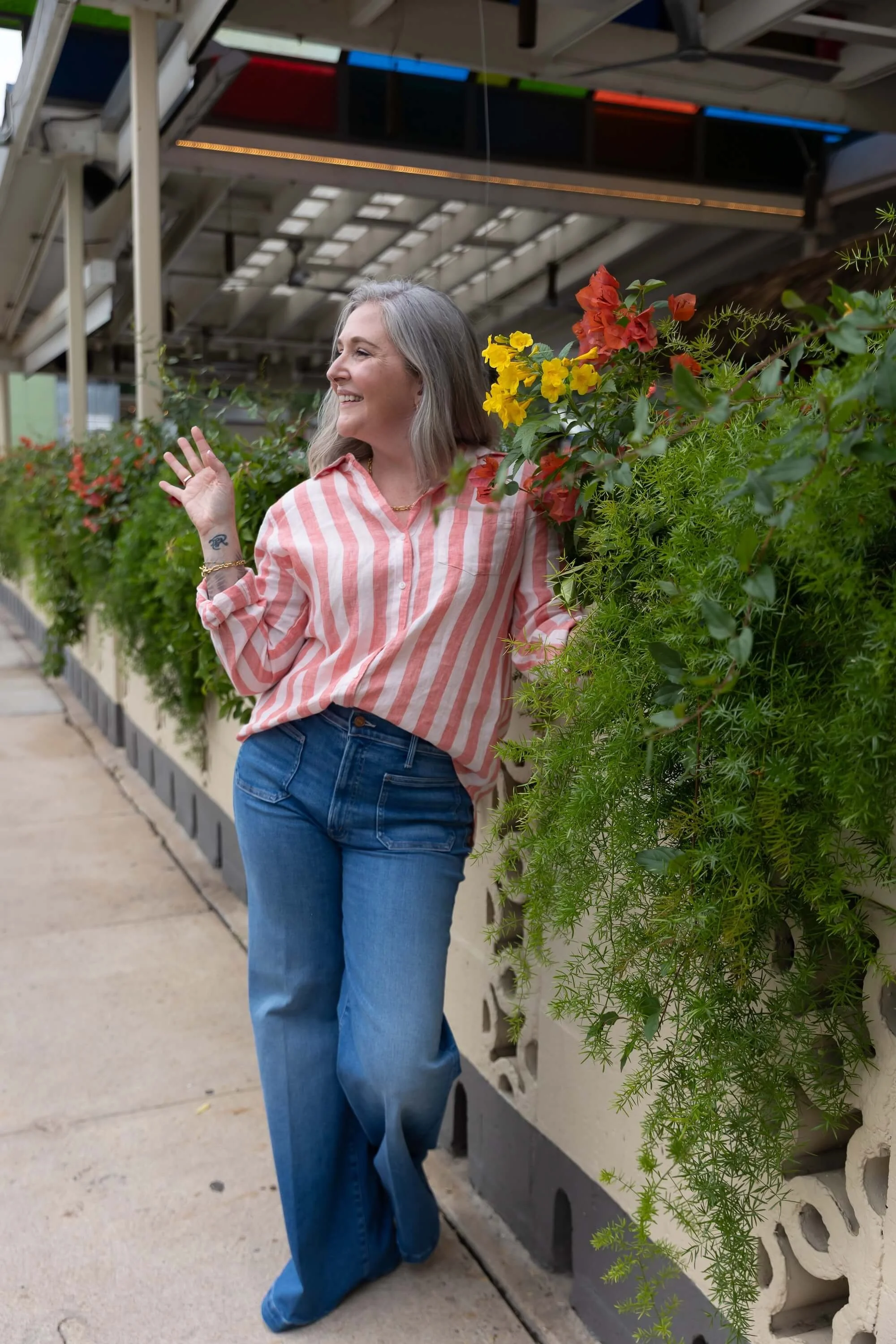 A woman with shoulder-length gray hair, wearing a pink and white striped shirt and blue jeans, smiling and waving while standing near a row of potted plants with orange and yellow flowers on a sidewalk under a metal shelter.