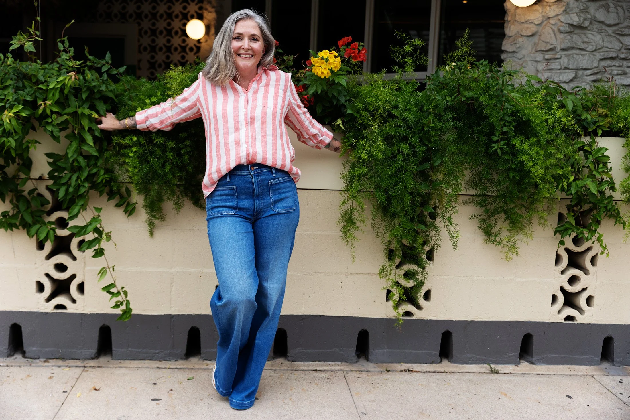 A woman with gray wavy hair smiling and wearing a pink and white striped shirt with blue jeans, leaning against a wall with green plants and red and yellow flowers, at dusk or early evening.