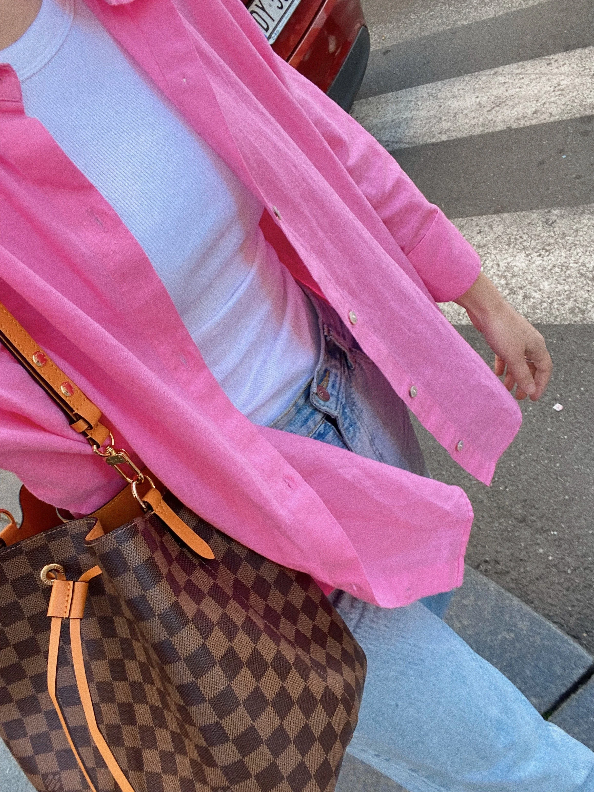 Close-up of a person's outfit showing a white ribbed top, a pink velvety over-shirt, light blue jeans, and a brown checkered handbag with tan leather accents, walking on a sidewalk with pedestrian crossing stripes.