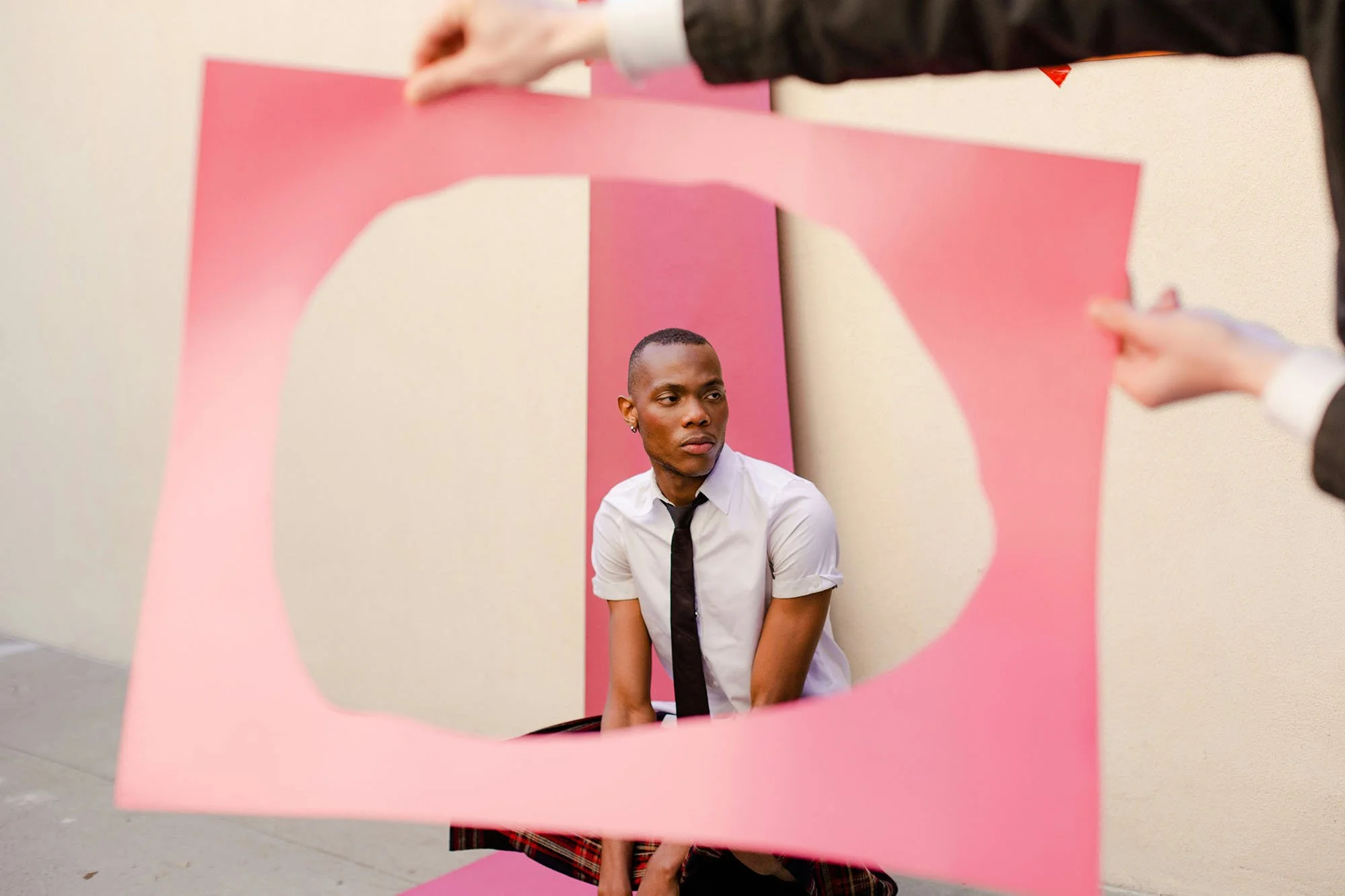A person with a short haircut and earrings looking to the side, wearing a white shirt and black tie, sitting in front of a pink backdrop with a cutout shape.
