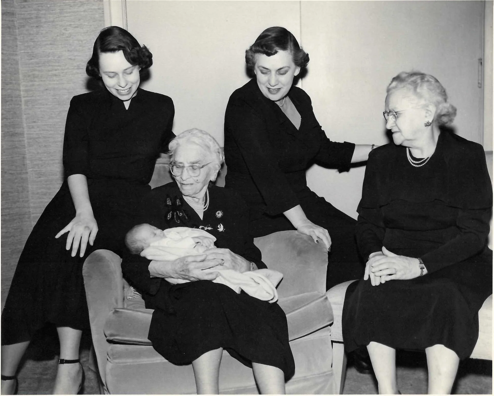 Four women, two elderly and two younger, are gathered around an elderly lady sitting on a chair, whose head is held gently by two of the women. They are in a room with light-colored walls, and the elderly woman is wearing glasses and a dark dress with a pattern. The two younger women are standing and leaning in, one on each side, with friendly smiles, while the elderly woman and the woman seated are looking at each other with a tender moment.